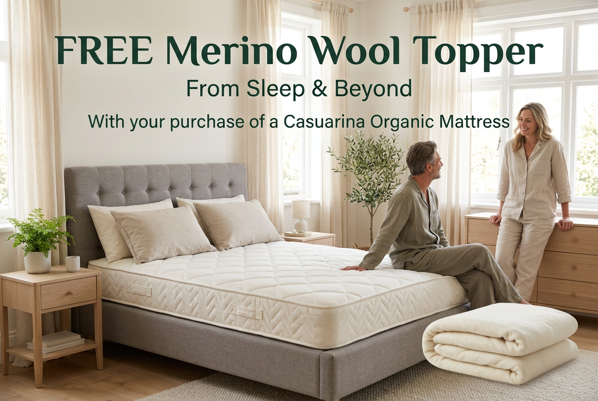 A bedroom scene showing a bed on a gray mattress base, with two people standing and sitting nearby. Large text at the top reads 'FREE Merino Wool Topper From Sleep & Beyond With your purchase of a Casuariana Organic Mattress