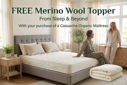 A bedroom scene showing a bed on a gray mattress base, with two people standing and sitting nearby. Large text at the top reads 'FREE Merino Wool Topper From Sleep & Beyond With your purchase of a Casuariana Organic Mattress